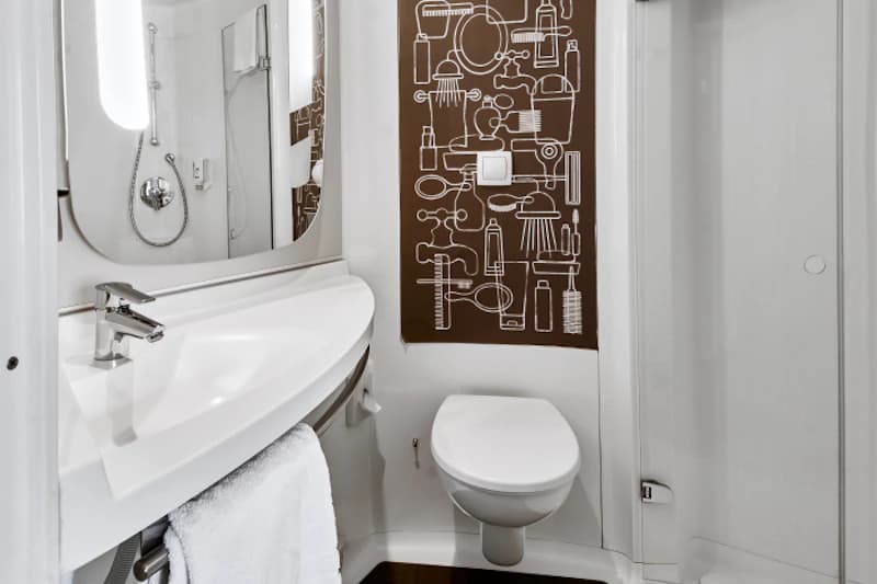 Kompaktes Hotelbad mit Waschbecken, rundem Spiegel, Toilette, Duschkabine und brauner Wand mit Pflegepiktogrammen.