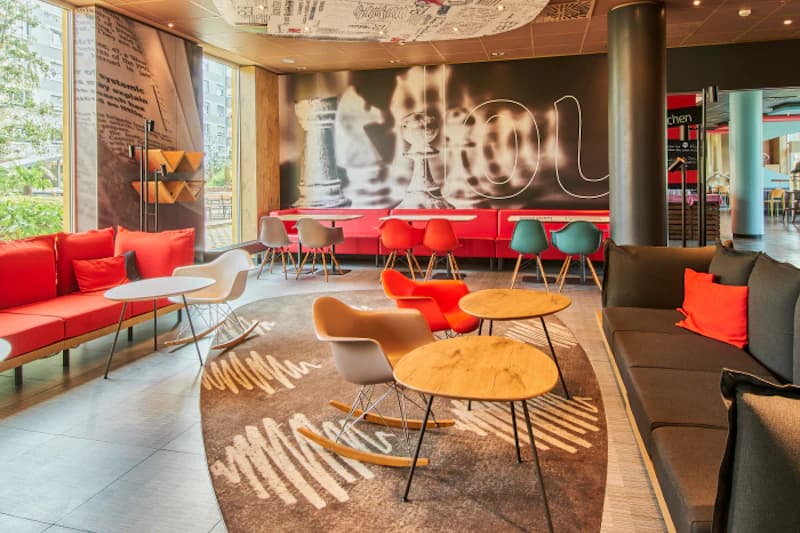 Hotellounge mit roten Sofas, bunten Stühlen, Holztischen und großem Wandbild mit Schachmotiven.