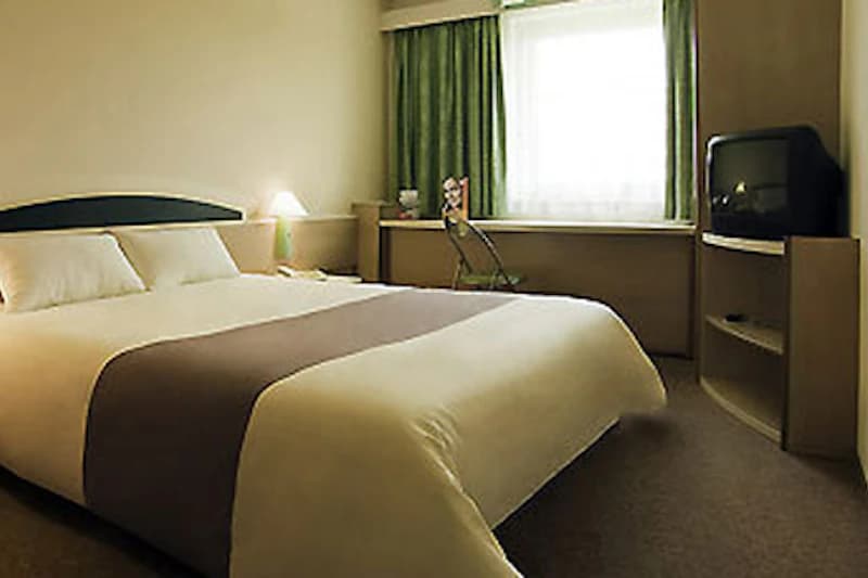 Doppelzimmer mit großem Bett, Nachttischlampe, Fenster mit grünen Vorhängen, Schreibtisch, Stuhl und Fernseher.