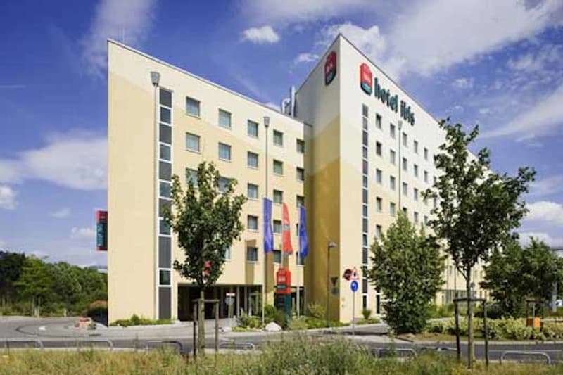 Mehrstöckiges helles Hotel mit B&B-Logo, Fahnen am Eingang und Bäumen im Vordergrund.