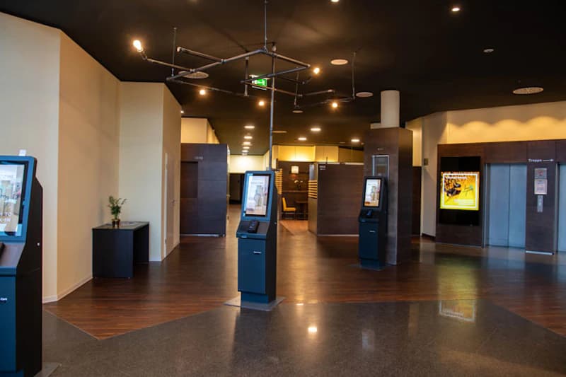 Hotellobby mit Selbstbedienungsautomaten, dunkler Decke, Holzfußboden und Aufzügen im Hintergrund.