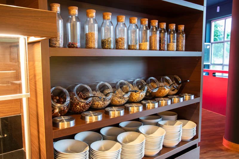 Offenes Regal mit Glasgefäßen gefüllt mit Müsli und Nüssen sowie gestapelten Schalen und Tellern.