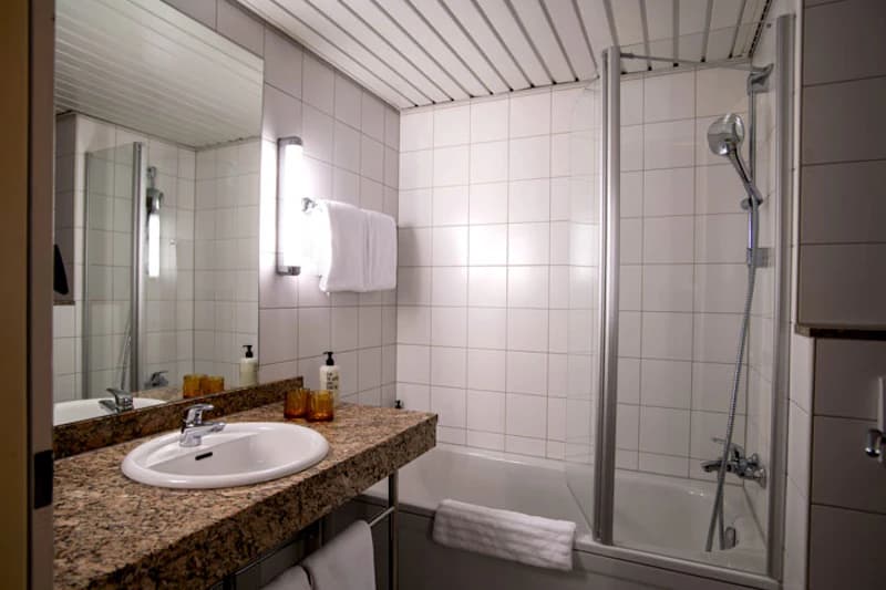 Weiß gefliestes Hotelbadezimmer mit Granitwaschbecken, beleuchtetem Spiegel, Handtuch und Badewanne mit Duschkopf.