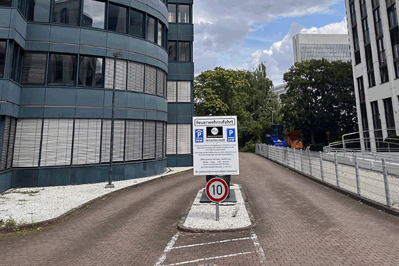 Zufahrt zwischen Bürogebäuden mit Feuerwehrzufahrt- und Parkschildern sowie Tempo-10-Schild.