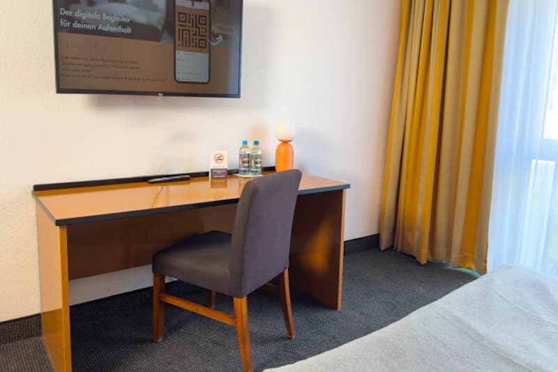 Schreibtisch mit Stuhl, Lampe, zwei Wasserflaschen und Wandfernseher mit QR-Code neben Vorhang.