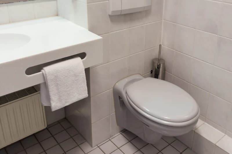 Wandhängendes WC mit geschlossenem Deckel, Waschbecken mit Handtuch, Kachelwände und Boden.