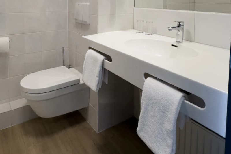 Weißes Waschbecken mit eingelassenem Handtuchfach, zwei Handtücher, Gläser und wandhängende Toilette.