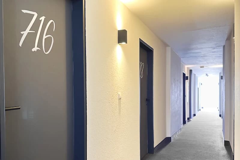 Flur mit grauen Zimmertüren, Zimmernummer 716 sichtbar, Wandlampen und Teppichboden bis zu hellem Ende.