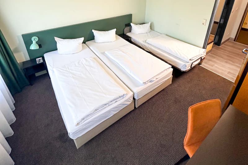 Hotelzimmer mit drei Einzelbetten an grünem Kopfteil, Nachttisch mit Lampe, oranger Stuhl und offener Tür.