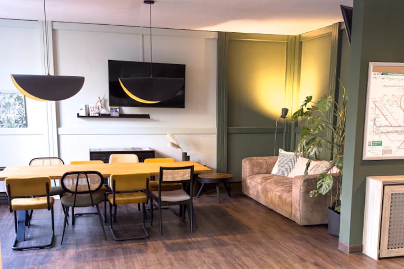 Gedeckter Gemeinschaftstisch mit Stühlen, Hängeleuchten, Fernseher und Sitzcouch in Hotelaufenthaltsraum.