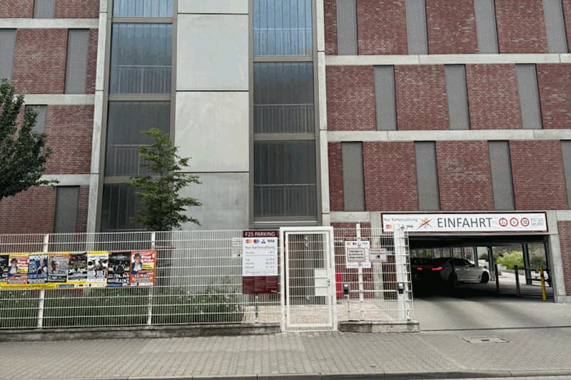 Mehrstöckiges Parkhaus mit Gitterzaun, Zugangstür, Einfahrtsschild und zwei einfahrenden Autos.