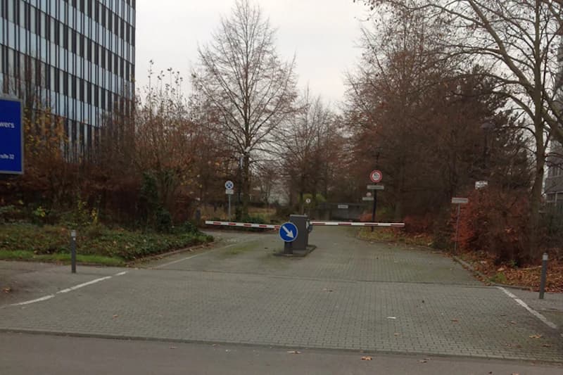 Zufahrt zur Tiefgarage mit geschlossenem Schranken und blauem Richtungsschild, Bäume und Bürogebäude im Hintergrund.