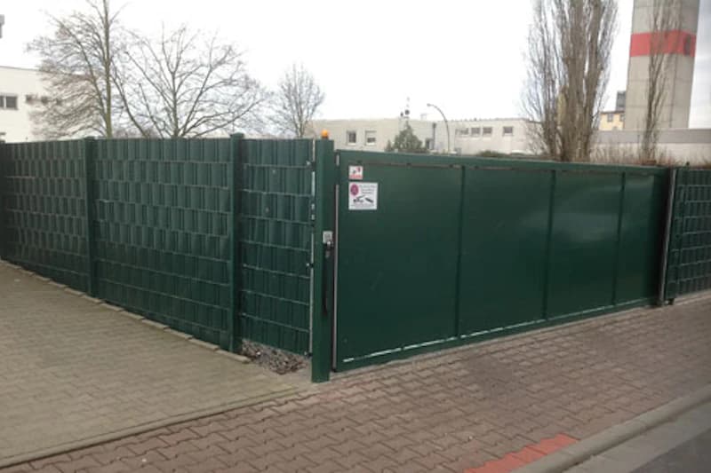 Geschlossenes grünes Schiebetor mit hoher Metallgitterumzäunung an gepflasterter Zufahrt.