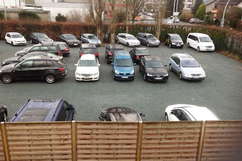 Schotterparkplatz mit mehreren geparkten Pkw, im Vordergrund Holzlattenzaun, im Hintergrund Hecke und Gebäude.