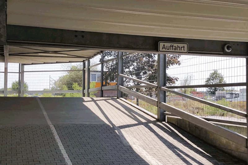 Innenrampe eines mehrstöckigen Parkhauses mit Auffahrt-Schild, Gittergeländer und diagonalen Sonnenschatten.