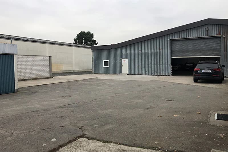 Gepflasterter Parkplatz vor grauer Metallhalle mit offenem Rolltor und schwarzem Auto am Eingang.