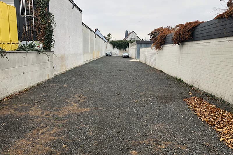 Schotterparkplatz zwischen hohen weißen Mauern, zwei geparkte Autos am Ende und Laubansammlung rechts.