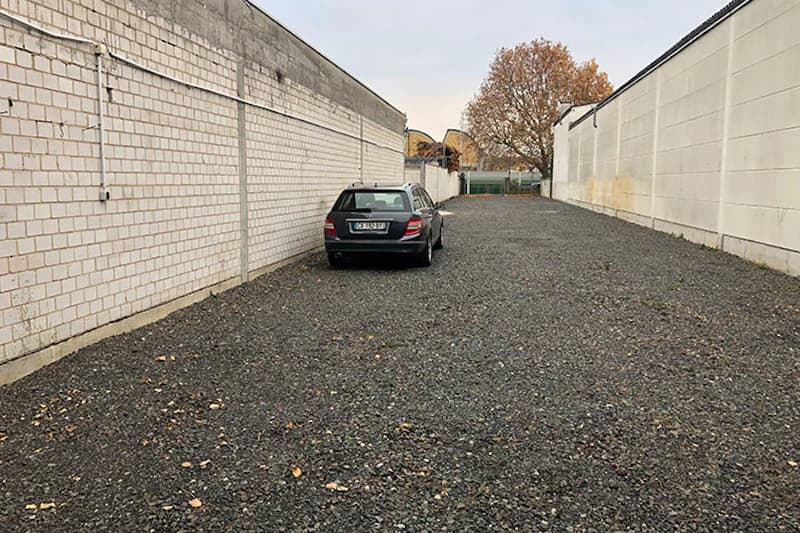 Schotterparkplatz zwischen hohen weißen Mauern mit einem geparkten dunklen Kombi und Baum am Ende.