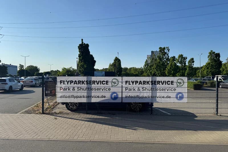 Gitterzaun mit Flyparkservice-Banner vor Gehweg und parkenden Autos auf einem Parkplatz.