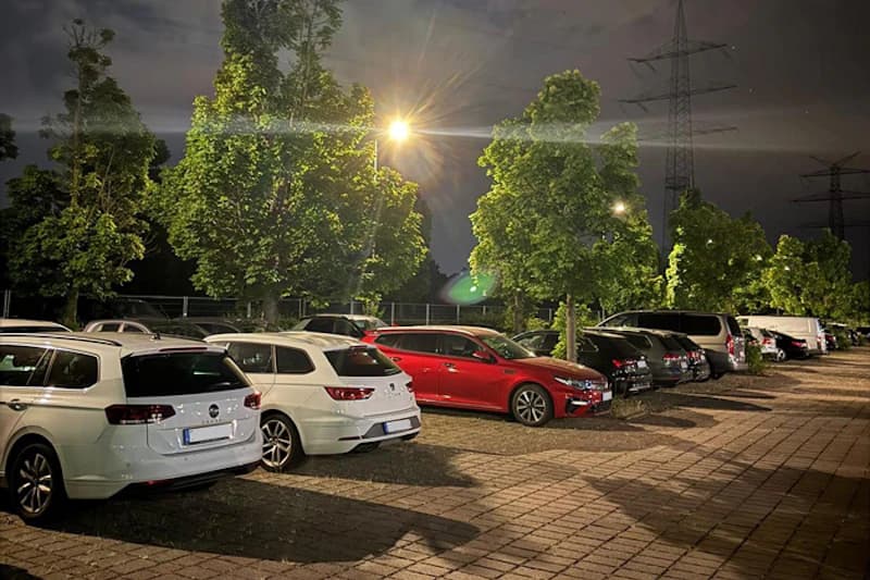 Reihe geparkter Autos auf beleuchtetem, eingezäuntem Parkplatz bei Nacht mit Bäumen und Strommasten.