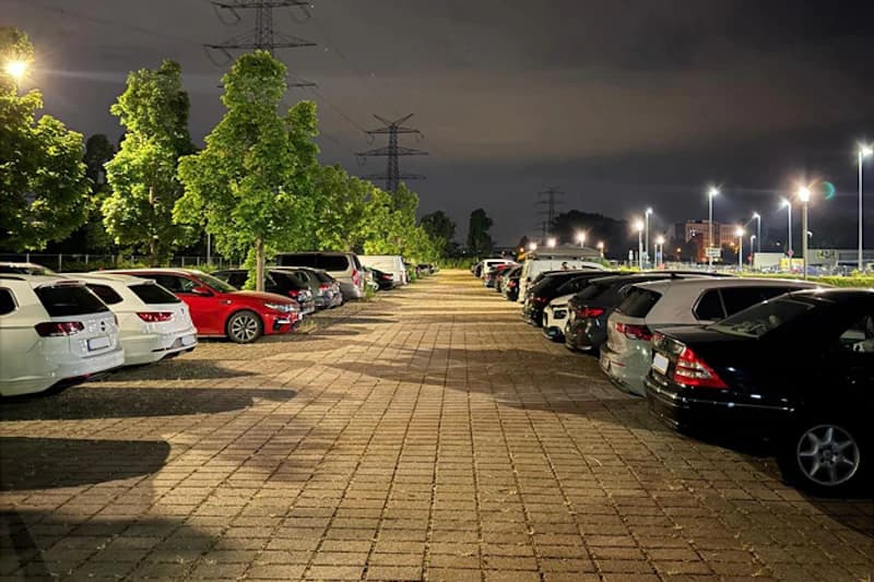 Beleuchteter Außenparkplatz bei Nacht mit gepflasterten Stellreihen, geparkten Autos, Bäumen und Hochspannungsmasten im Hintergrund.