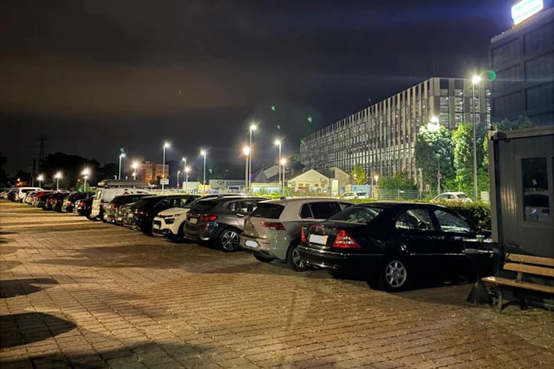 Geparkte Autos in einer Reihe auf beleuchtetem Außenparkplatz neben mehrstöckigem Gebäude bei Nacht.
