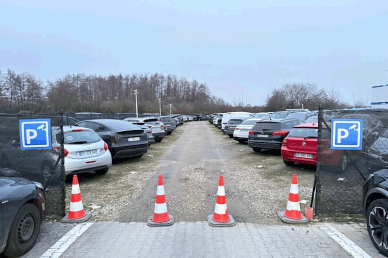 Einfahrt zu umzäuntem Parkplatz mit drei Leitkegeln, Reihen geparkter Autos und blauen Parkschilden.