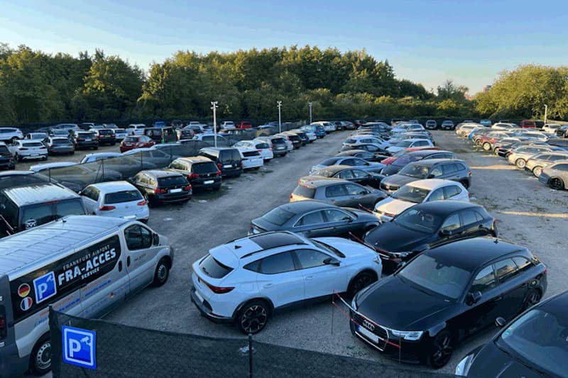 Umzäunter Parkhof mit dicht geparkten Autos, beleuchteten Pfosten, FRA-ACCES Shuttlebus und Baumreihe im Hintergrund.