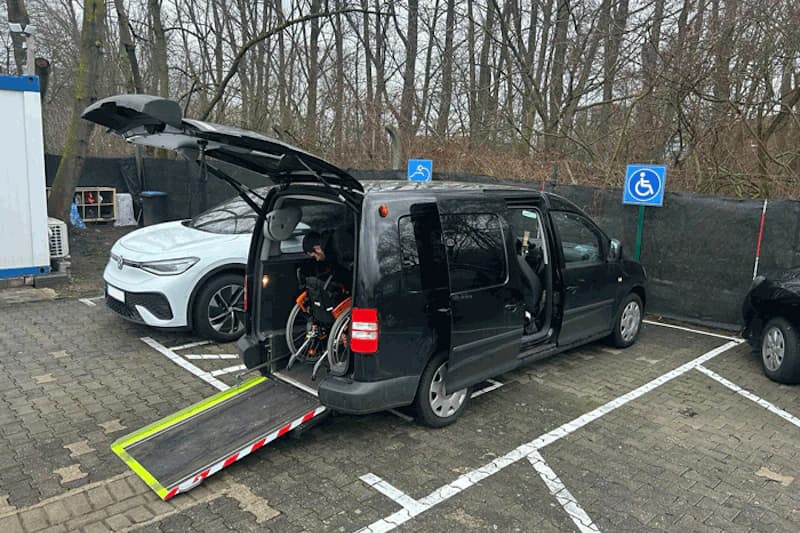 Schwarzer behindertengerechter Kleinbus mit ausgeklappter Rampe und Rollstuhl auf Parkplatz am Flughafen.