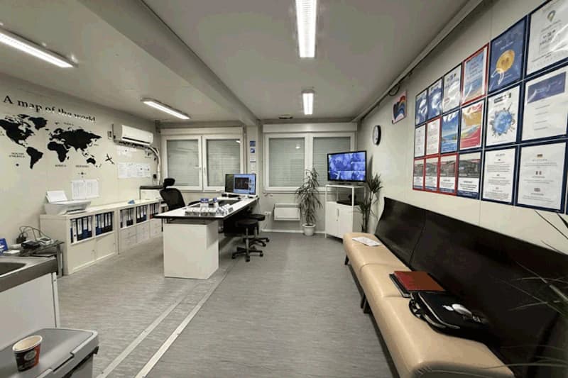 Büro mit Schreibtisch, mehreren Monitoren und Überwachungsbildschirm, Weltkarte an der Wand, Zertifikate und Sitzbank.