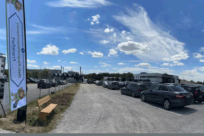 Kiesweg zwischen Reihen geparkter Autos, Metallzaun mit Fahne und Holzbank unter blauem Himmel.