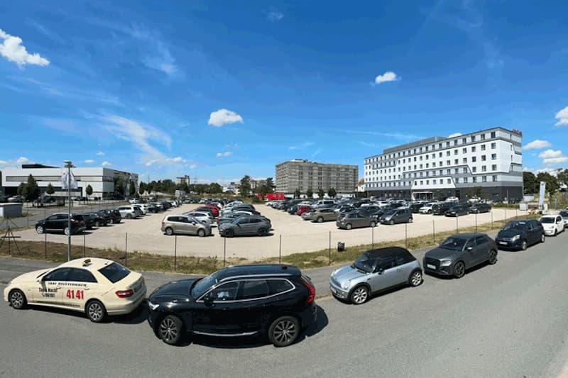 Gitterzaun umgebenes Schotterparkfeld mit geparkten Autos; großes Hotelgebäude rechts, Taxi in Vordergrund.