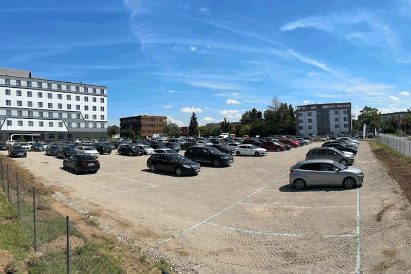 Großer geschotterter Parkplatz mit zahlreichen geparkten Autos, Umzäunung und Gebäuden im Hintergrund unter blauem Himmel.