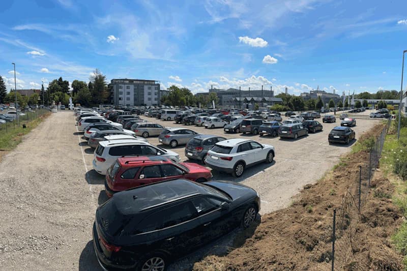 Großer offener Schotterparkplatz mit zahlreichen geparkten Autos, Umzäunung rechts und Gebäuden im Hintergrund.
