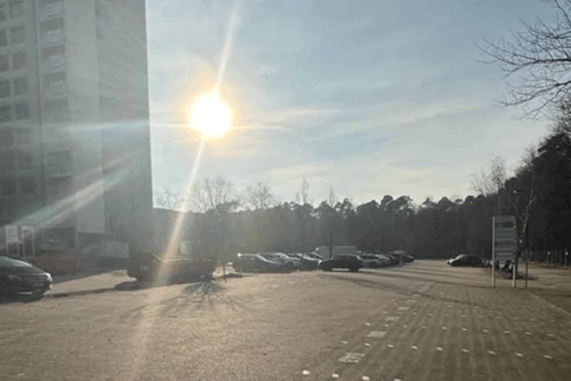 Parkplatz mit mehreren geparkten Autos, gepflasterte Fläche, tiefstehende Sonne und Gebäude links.