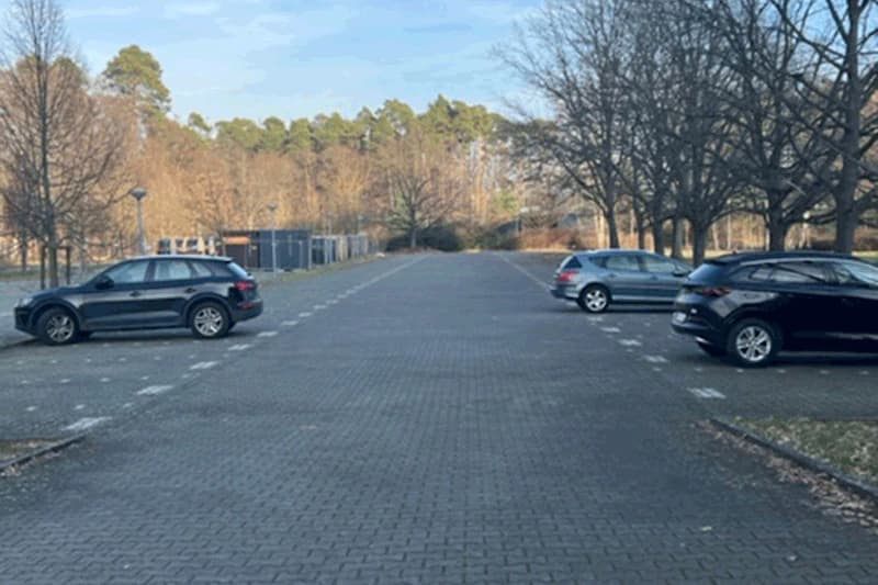 Gepflasterter Parkplatz mit einzelnen Autos, Baumreihe rechts und Containern am hinteren Rand.