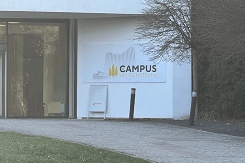 Eingang mit Glastür, Schild Campus Heusenstamm, Aufsteller mit Pfeil und zwei Poller am gepflasterten Weg.