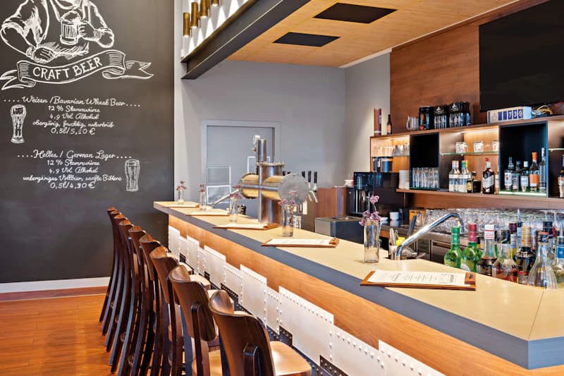 Hotelbar mit Tresen, Zapfanlage, hohen Barhockern und Regal mit Flaschen.