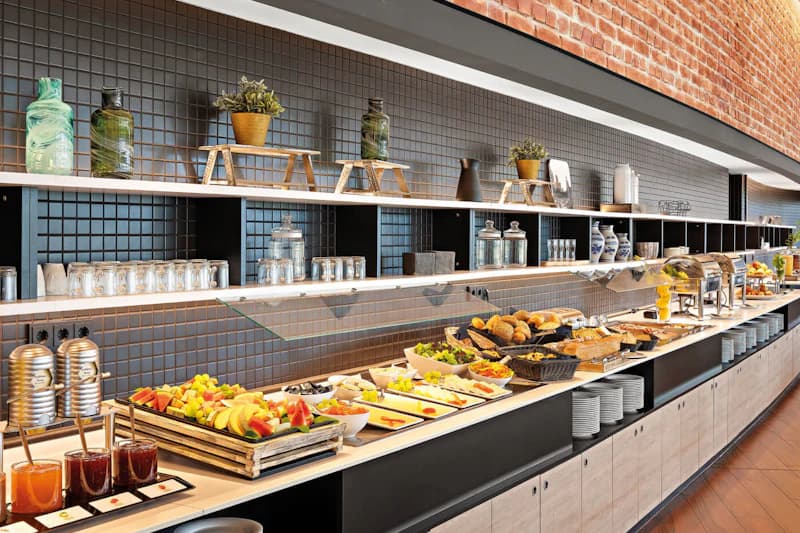 Buffet mit Obst, Aufschnitt, Gebäck, Schalen und Tellern vor dunkler Kachelwand mit Regalen und Vasen.