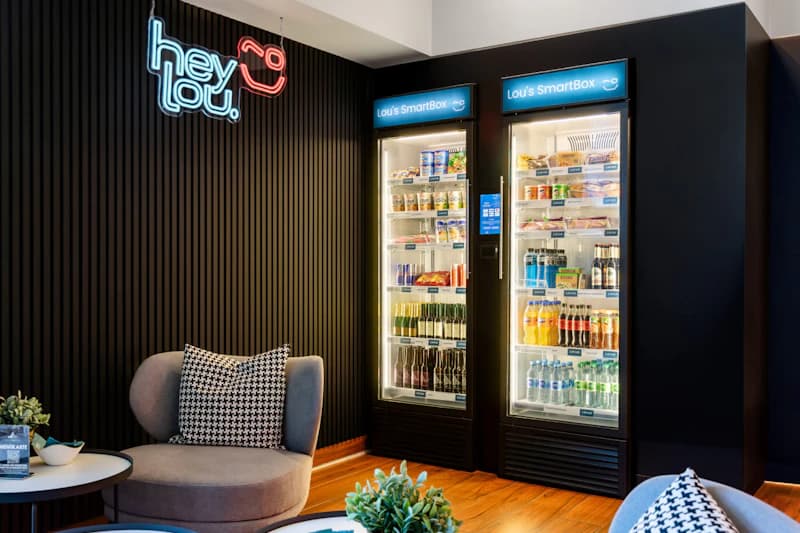 Zwei beleuchtete Verkaufskühlschränke neben Sitzbereich mit neon Hey Lou Schriftzug und Pflanzen.