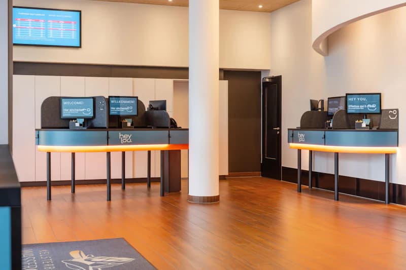 Hotelempfang mit drei Selbstbedienungsterminals, runder Säule, Monitortafel und Holzfußboden.