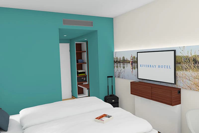 Hotelzimmer mit Doppelbett, türkiser Akzentwand, Wandfernseher, offenem Kleiderschrank und Rollkoffer.