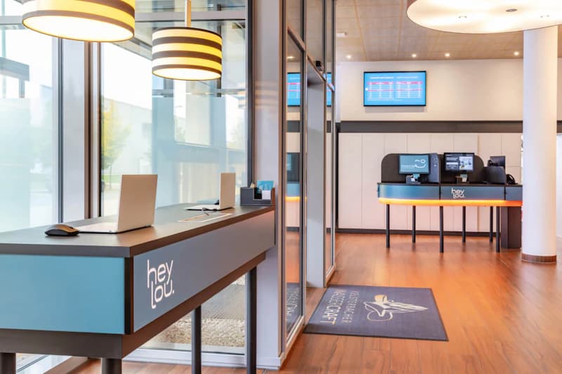 Hotelempfang mit Tresen, Selbstbedienungsterminals, Laptops und Wandmonitoren im Hey Lou Hotel am Flughafen Frankfurt.