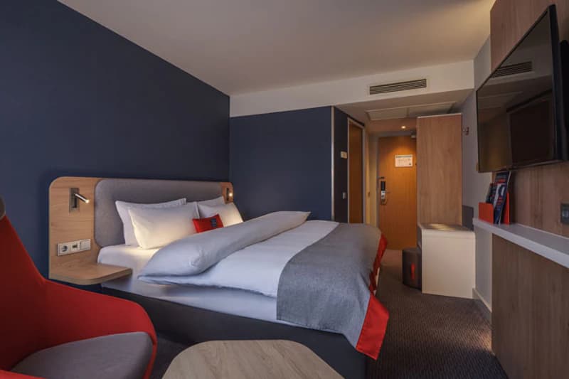 Hotelzimmer mit großem Doppelbett, dunkelblauer Wand, rotem Sessel, Wandfernseher und geöffneter Zimmertür.