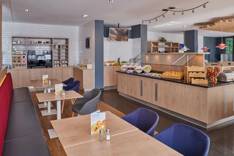 Frühstücksraum mit Buffet, Schalen mit Obst, Brötchen und Brezeln, Kaffeeautomat sowie Tischen und Stühlen.