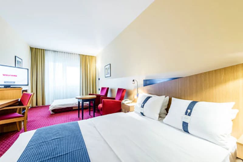 Helles Hotelzimmer mit Doppelbett, Nachttisch, zwei roten Sesseln, Tisch, Schreibtisch, Fernseher und Fenster mit Vorhängen.