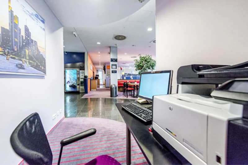 Arbeitsplatz mit Computer, Tastatur und Drucker in einer Hotellobby, Sitzbereich und Getränkeautomat im Hintergrund.