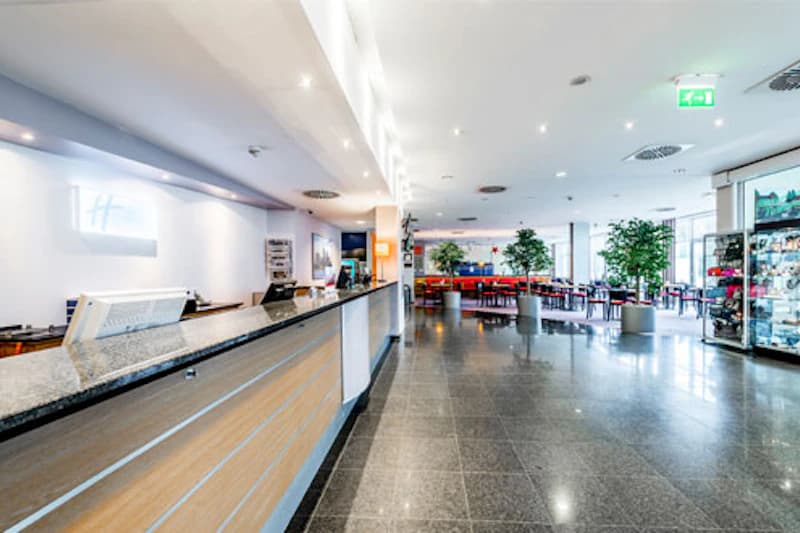 Rezeption mit langem Tresen links, offene Hotellobby mit Sitzgruppen, Pflanzen und Vitrine am Flughafen Frankfurt.