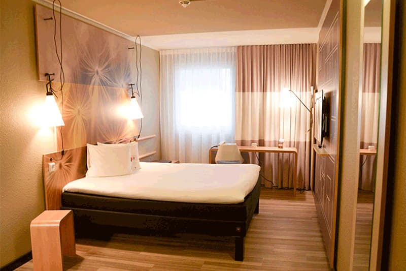 Hotelzimmer mit Bett, zwei hängenden Lampen, gemustertem Kopfteil, Fenster, Schreibtisch und Wandspiegel.