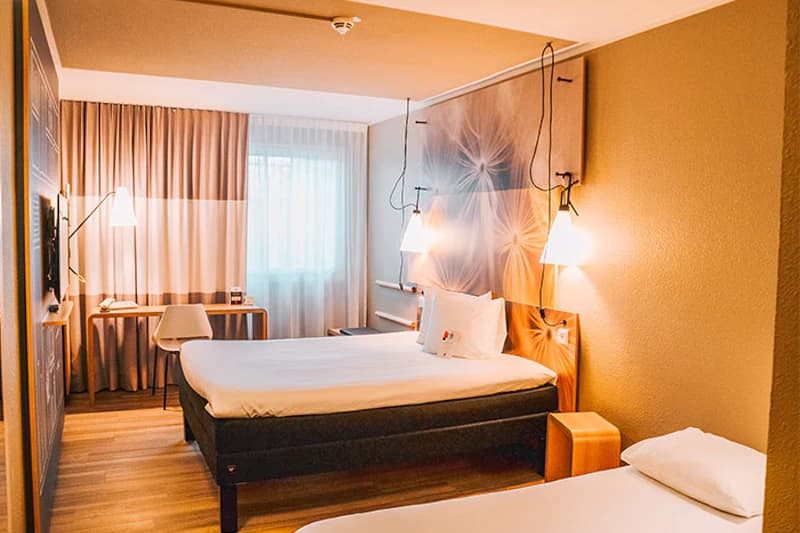 Hotelzimmer mit Doppelbett und Einzelbett, hängenden Nachttischlampen, Schreibtisch am Fenster und Vorhängen.